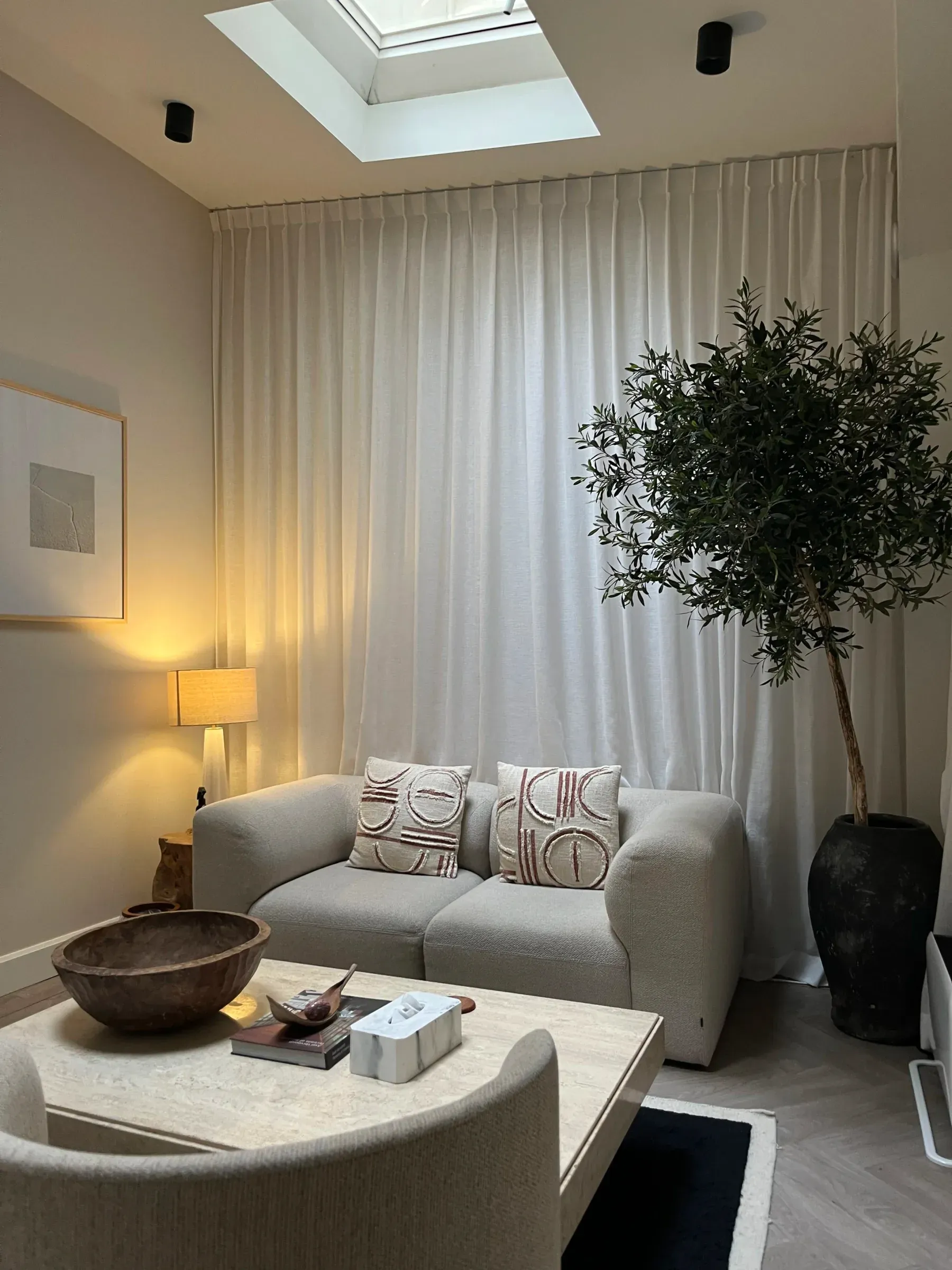 The therapy room at Keizersgracht: a soft grey sofa beneath a skylight, linen curtains, a warm lamp and an olive tree.